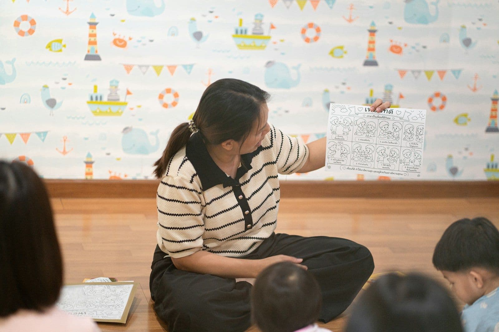 A teacher holding up a worksheet for a group of small children in a room with nautical wallpaper