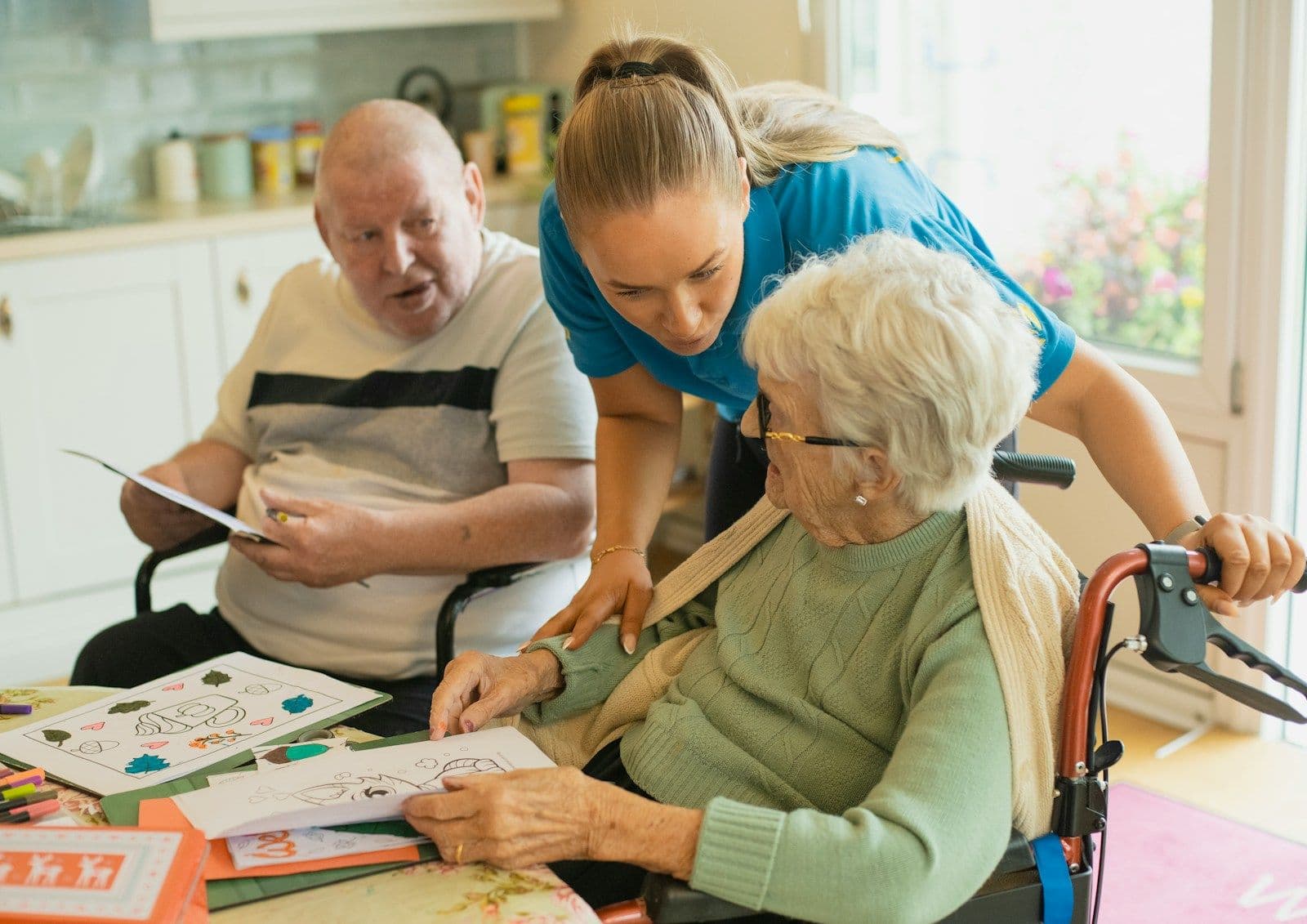 A caregiver leaning in to help an older couple with a colouring page at their kitchen table